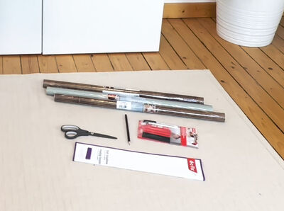 Die Bestandteile des Kreativ-Kits liegen auf einem Teppich. Zwei Klebefolienrollen, die Faltschablone, eine Schere, Lineal sowie das d-c-fix Verarbeitungsset mit Rakel.